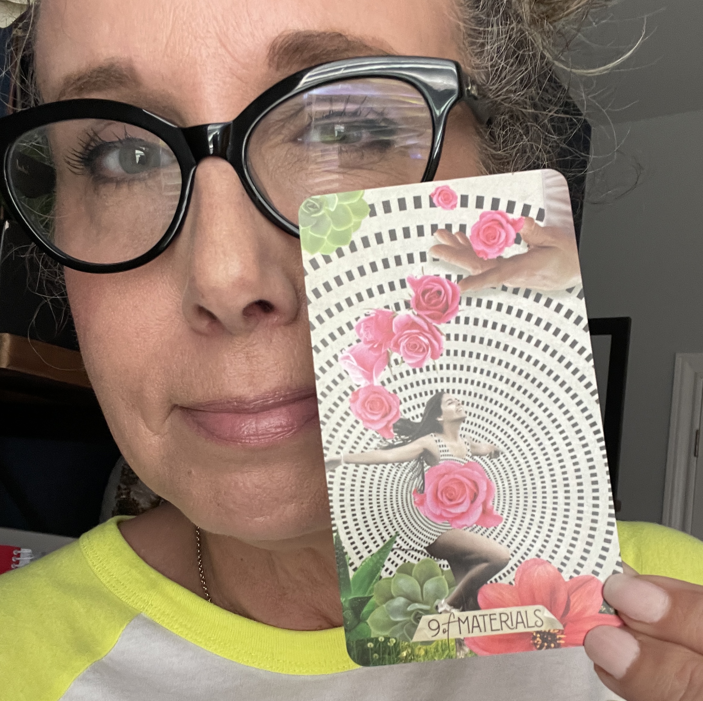 Allison holding the 9 of Materials card from Chris Anne's Must Tarot Deck. A woman in a black and white spiral with arms open wide and blooms of pink roses on her body and trailing to the sky.