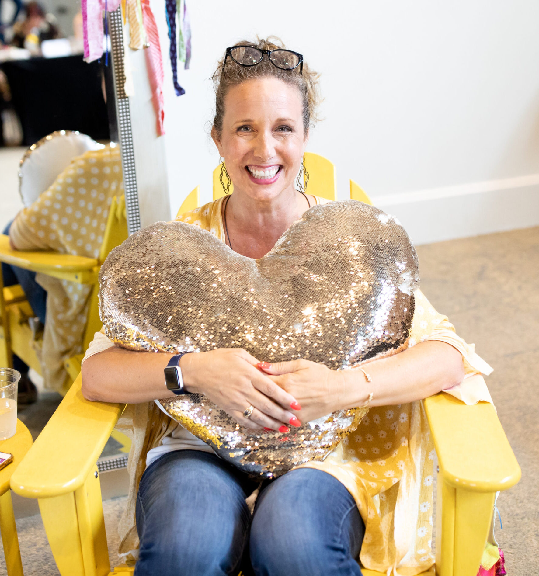 Allison sitting in a yellow chair with a big smile holding a giant gold heart-shaped pillow in her arms.
