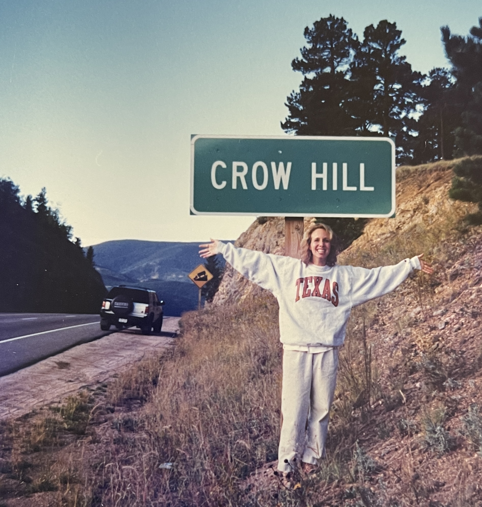 Allison standing on the side of hwy 285 in colorado with her arms open wide.  Just behind her is a sign that says CROW HILL and beyond that her 1995 Isuzu rodeo is parked on the edge of the road. 