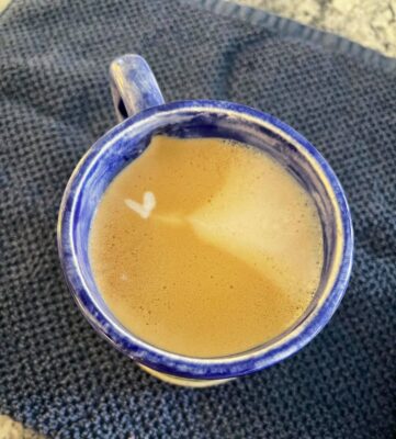 A blue cup of coffee with cream that has a heart shaped white bubble in it.